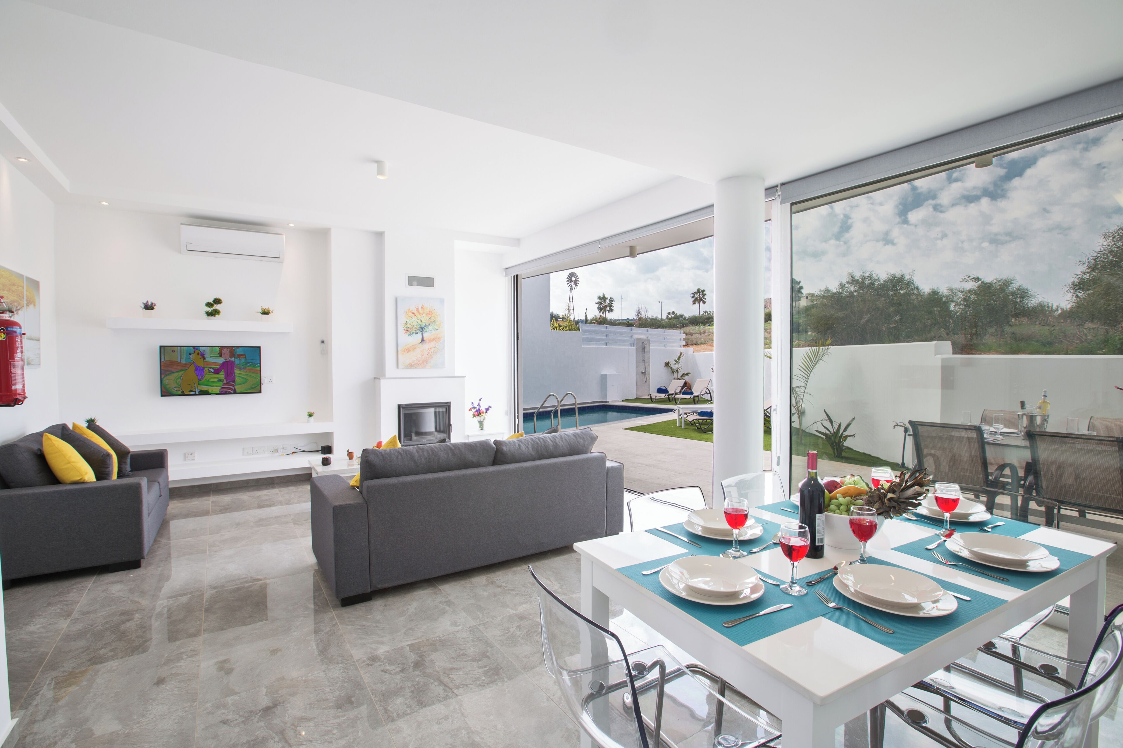 Uma sala de estar moderna com um sofá cinzento, uma mesa de jantar posta para quatro pessoas e uma grande janela com vista para uma piscina e área externa.