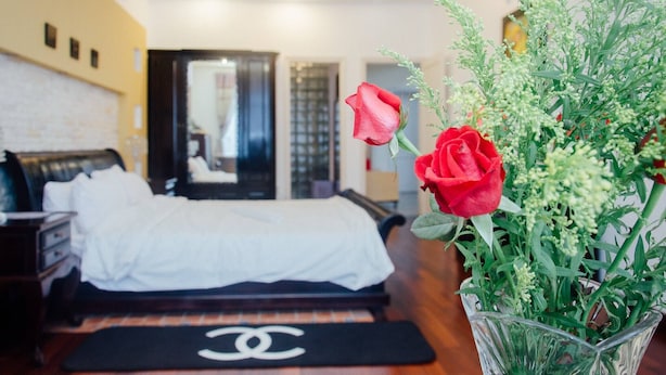 A hotel room with a bed, a Chanel-branded mat, and a vase with red roses and greenery.