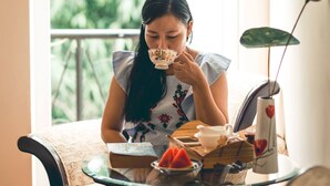 A woman sitting at a table with a floral dress, a teapot, and a vase with a flower. - Hillside Homestay Hue - Silk Room (Hue)