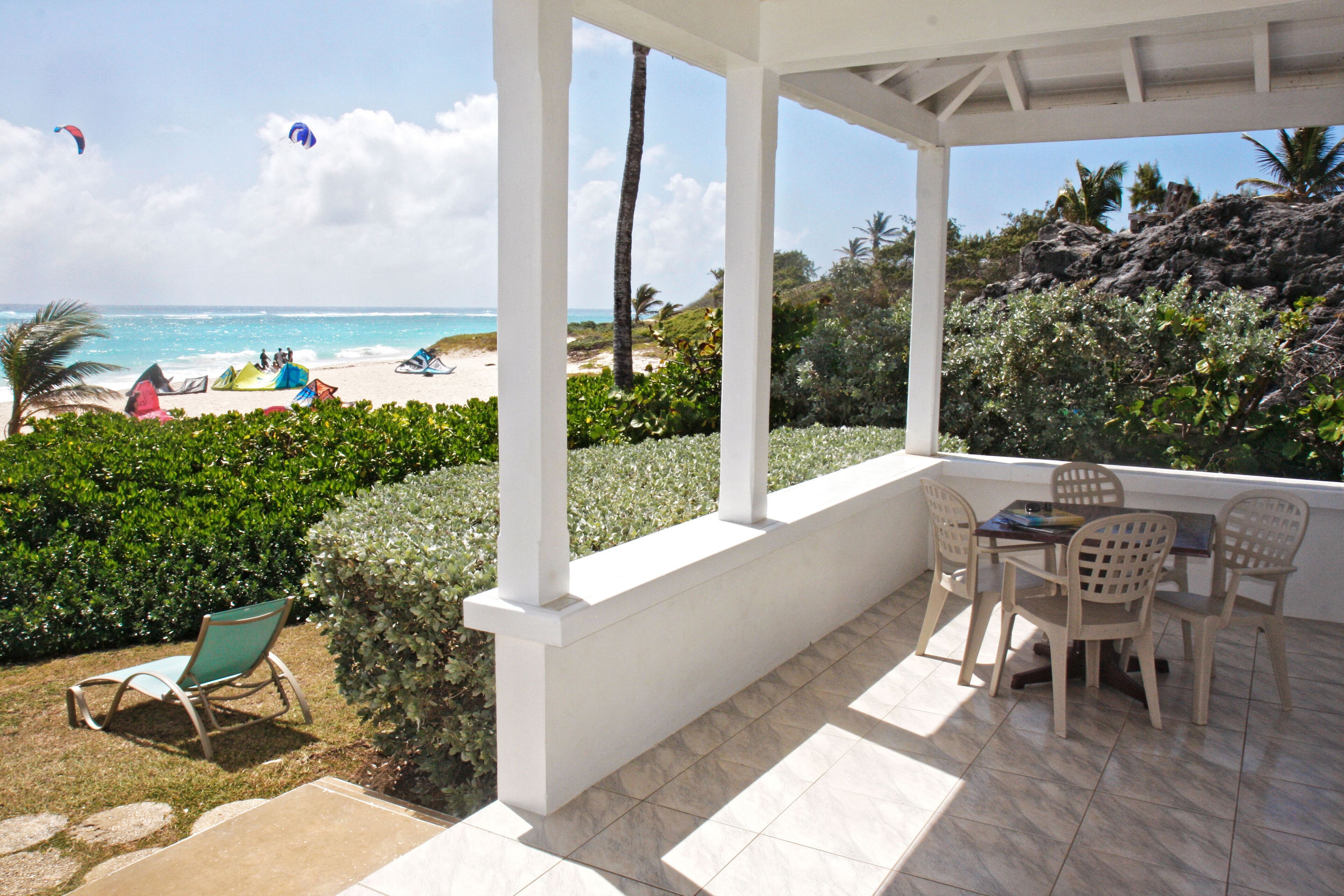 A covered patio with a table and chairs, overlooking a beach with tents and people.