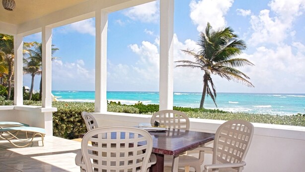 A covered outdoor seating area with a table and chairs, overlooking a beach with palm trees and the ocean. - Silver Sands #1 - by Zenbreak (Silver Sands)
