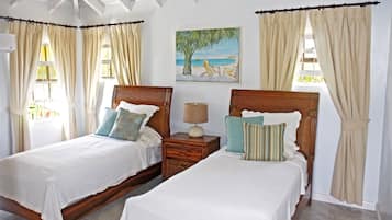 A bedroom with two wooden beds, beige curtains, a beach-themed painting, and a small bedside table with a lamp.