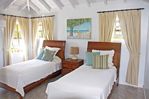 A bedroom with two wooden beds, beige curtains, a beach-themed painting, and a small bedside table with a lamp. - Silver Sands #2 - by Zenbreak (Silver Sands)