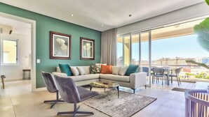 A modern living room with a grey sofa, a black chair, and a coffee table. There are two framed pictures on the wall, a large window with a view of buildings, and a balcony with outdoor furniture. - Luxury Three Bedroom Apartment - Fully Furnished and Equipped (Cape Town)