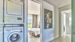 A modern hotel room with a Bosch washing machine, a bed with a bench, a large window, and a tropical-themed painting. - Luxury Three Bedroom Apartment - Fully Furnished and Equipped (Cape Town)