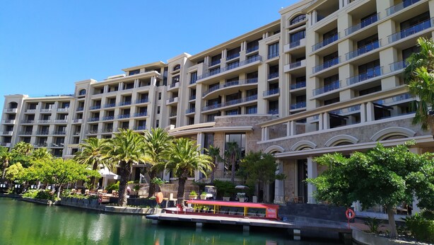 A multi-story hotel with balconies, a water body in front, and a small boat dock. - Luxury Three Bedroom Apartment - Fully Furnished and Equipped (Cape Town)