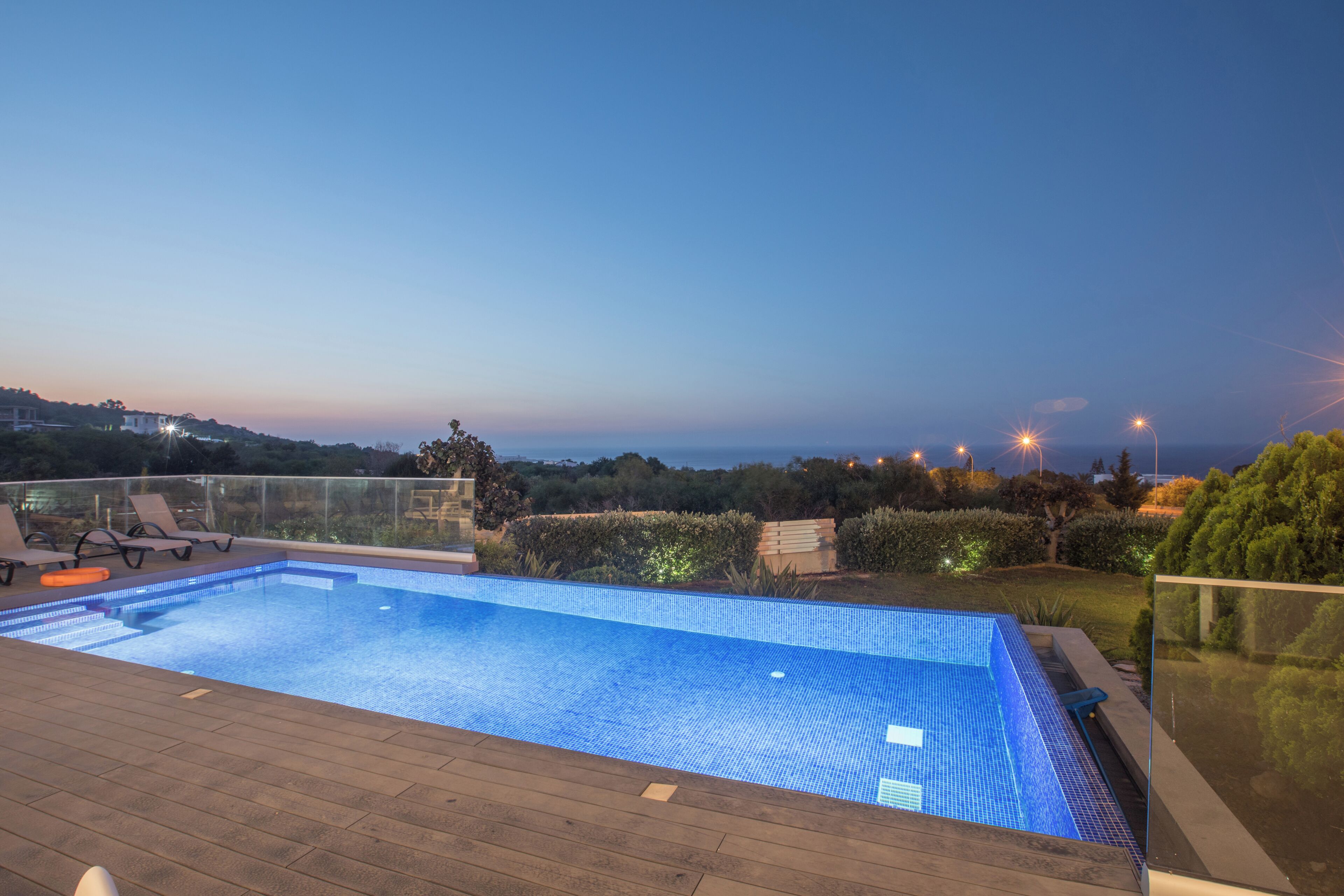 A swimming pool with a wooden deck, surrounded by a glass railing and a view of the landscape.