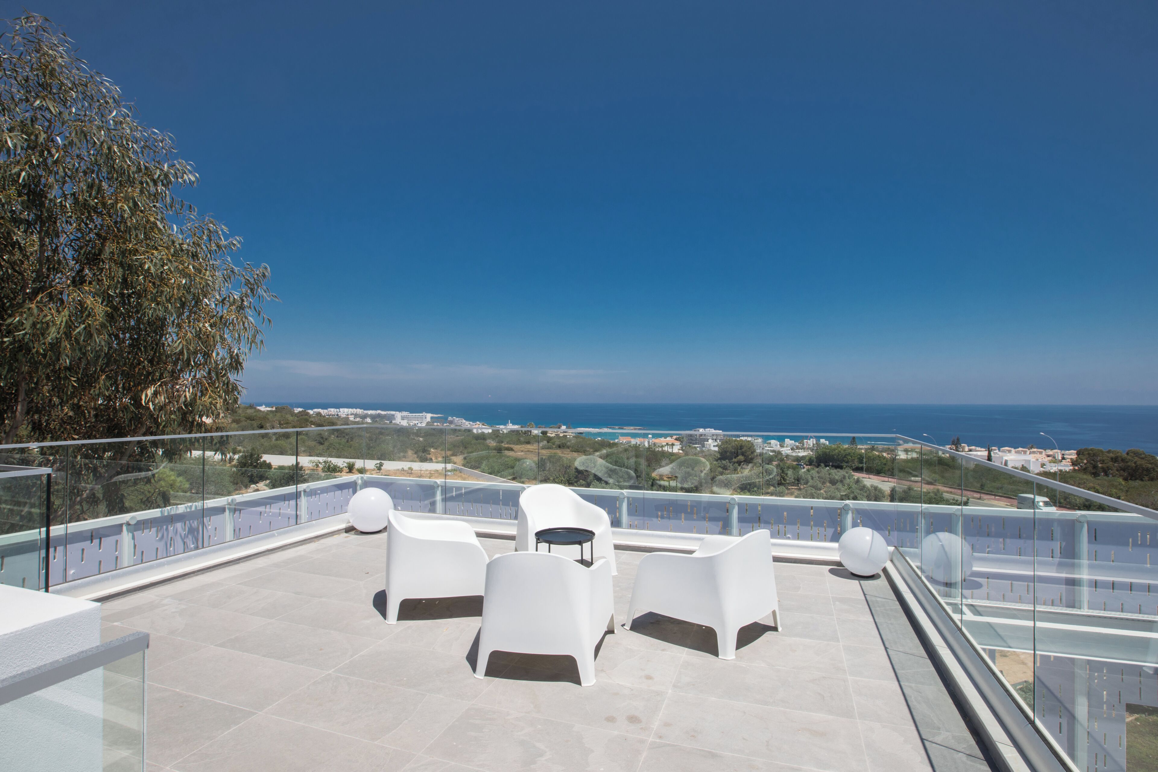 A rooftop terrace with white plastic chairs and a glass balustrade overlooking a coastal landscape.
