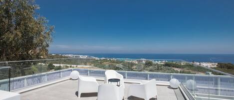A rooftop terrace with white plastic chairs and a glass balustrade overlooking a coastal landscape.