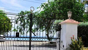 Une entrée fermée par un portail en fer forgé décoratif, un pilier blanc orné d’une fleur rouge, et une vue sur la mer que l’on aperçoit à travers le portail.