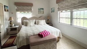 Une chambre à coucher comprenant un lit, des tables de chevet, une fenêtre ornée de barreaux en fer décoratifs et un ventilateur de plafond.