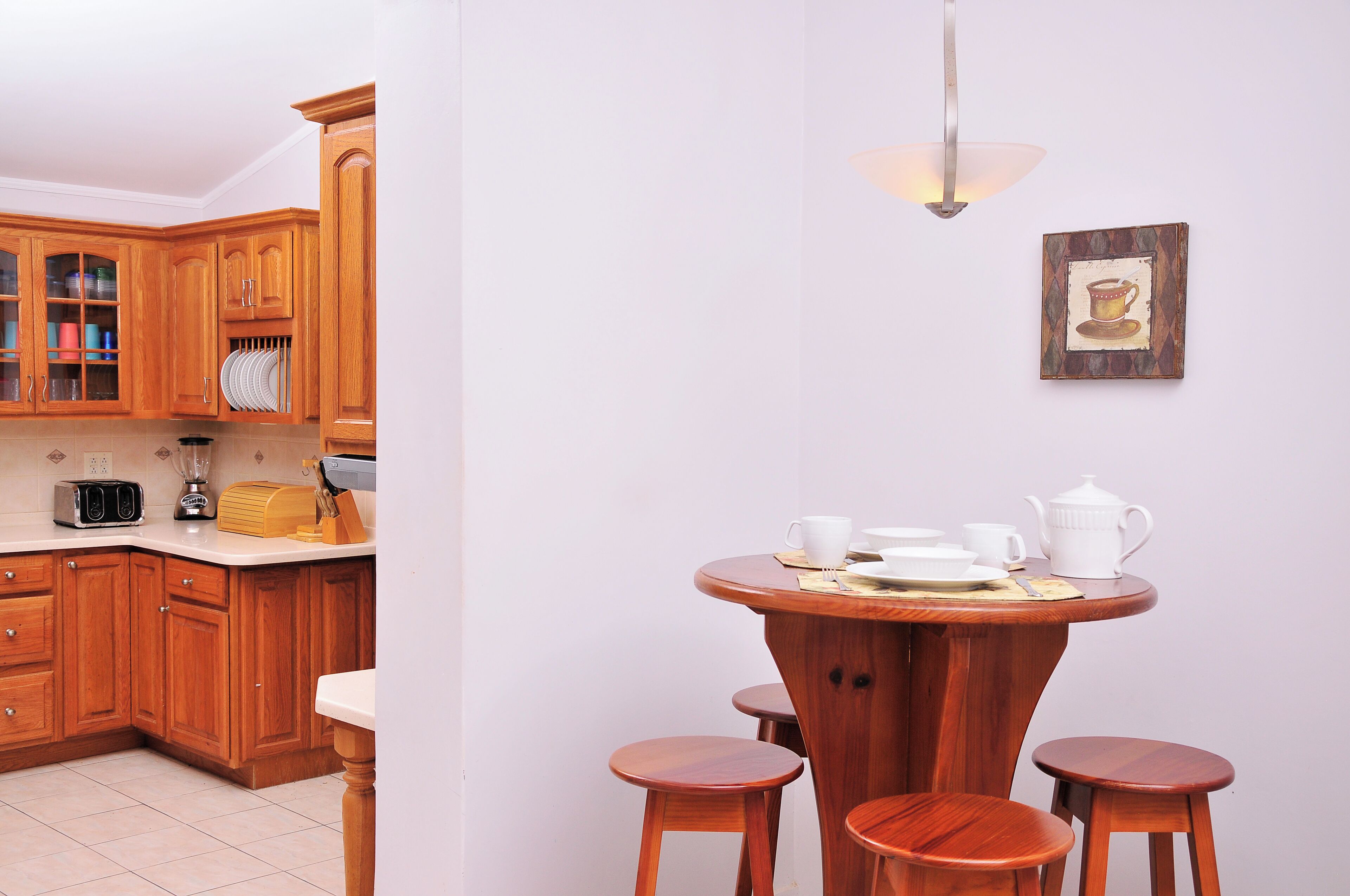 Une cuisine avec des meubles de rangement en bois, un plan de travail sur lequel se trouvent un grille-pain, un mixeur et une machine à café. On y trouve également une table à manger avec quatre tabourets et un luminaire fixé au mur.