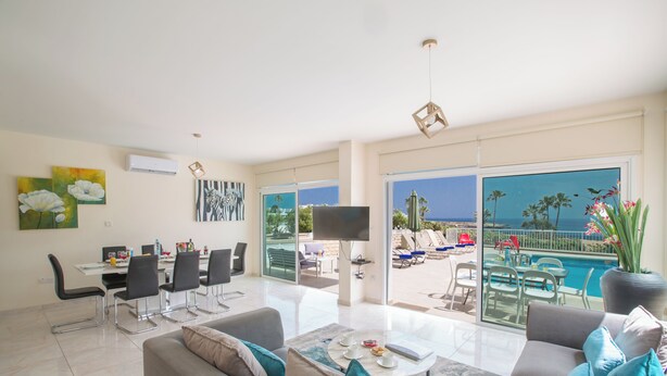 A spacious living room with a sofa, coffee table, and dining area. A large TV is mounted on the wall, and there is a balcony with a pool view. - Protaras Mesogeios Villa Me16 Beachfront Villa in Protaras Center (Paralimni)