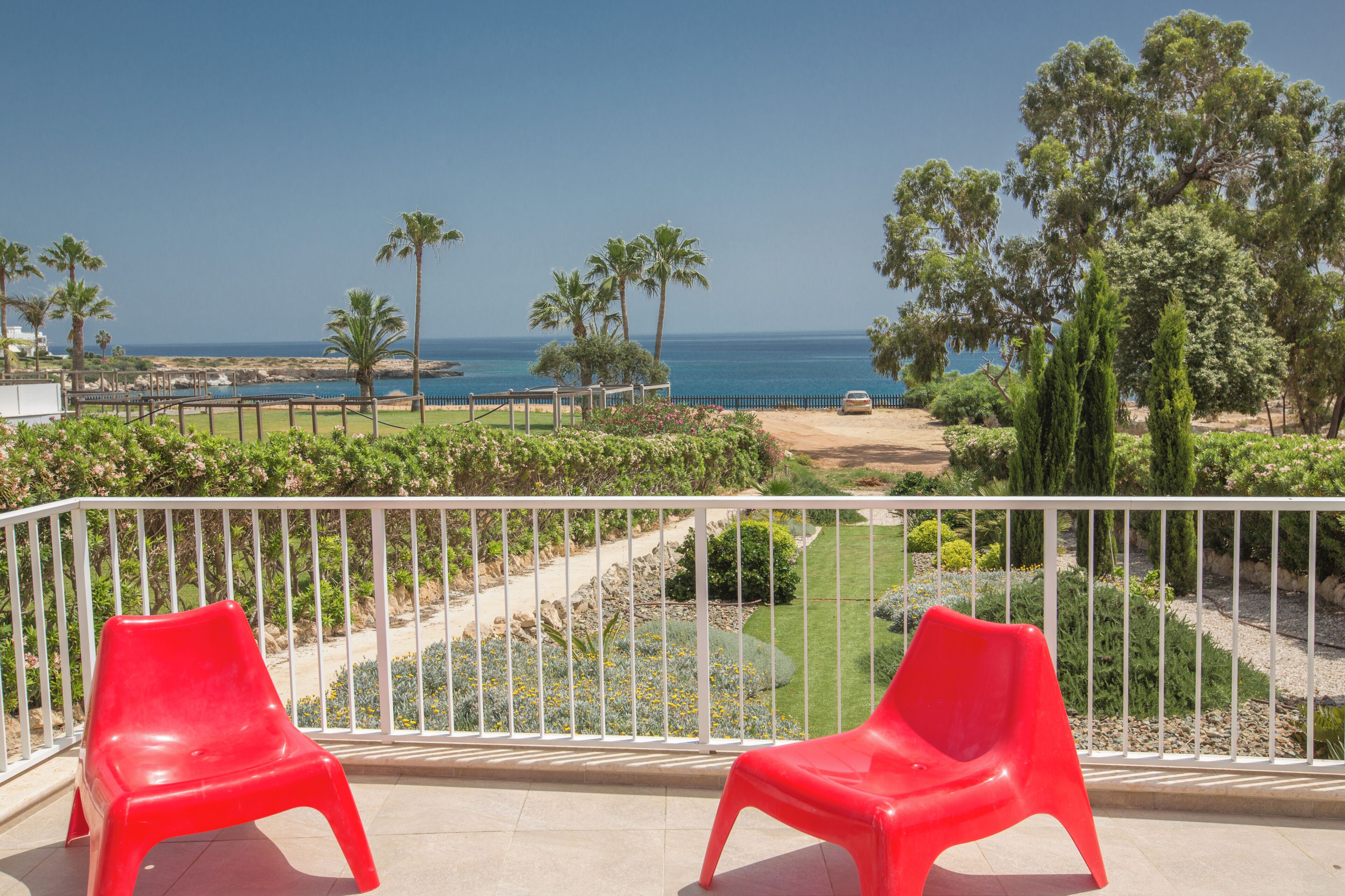 Un balcon avec deux chaises en plastique rouge, une balustrade blanche et une vue sur la mer et la végétation.