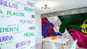 Un espace repas moderne avec une table et des chaises blanches, une fresque colorée sur le mur et un luminaire au plafond.
