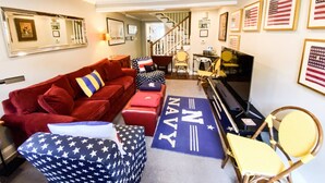 A living room with a red sofa, a piano, and a rug with 'Navy' written on it. There are framed pictures on the wall and a staircase in the background. - Go Navy! Steps to USNA, City Dock & Street Parking (Annapolis)