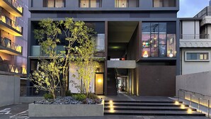 Un edificio moderno con entrada central, un pequeño jardín con árboles y una escalera que conduce a la entrada.