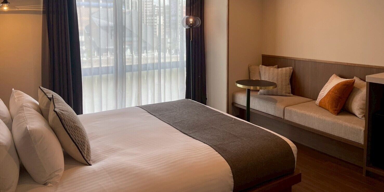 A hotel room with a large bed, a sofa, a small round table, and a view through sheer curtains.