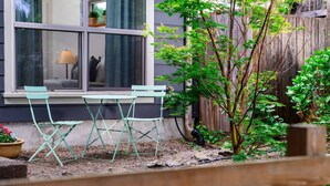 Un pequeño espacio al aire libre con dos sillas verdes y una mesa, una ventana con vista a una casa y una valla de madera.