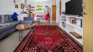 Una sala de estar moderna con un sofá, una mesa de centro y un tapete rojo estampado. Hay un televisor en la pared y una estantería con libros.