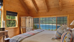 A wooden cabin bedroom with a large bed, a wooden wardrobe, and a view of a lake and forest through large windows.