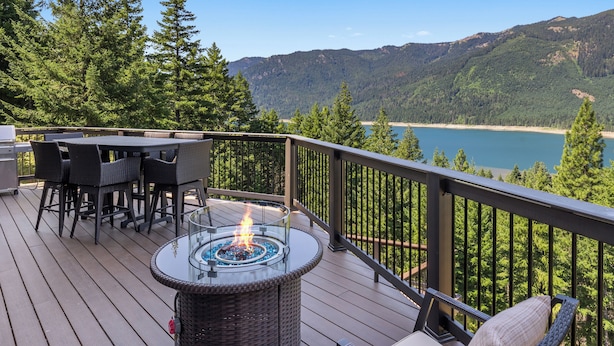 A deck with a glass-top fire pit, wicker furniture, and a view of a lake and mountains.