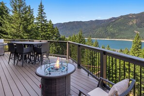 Une terrasse dotée d’un foyer extérieur avec un plateau en verre, de meubles en osier et offrant une vue sur un lac et des montagnes.