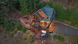 A large, multi-story lodge with a deck and hot tub, surrounded by dense forest.