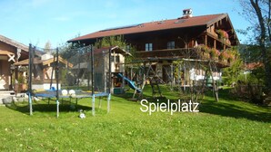 Un patio de juegos con un trampolín y toboganes, rodeado de un césped cuidado y una casita de madera con techo rojo.