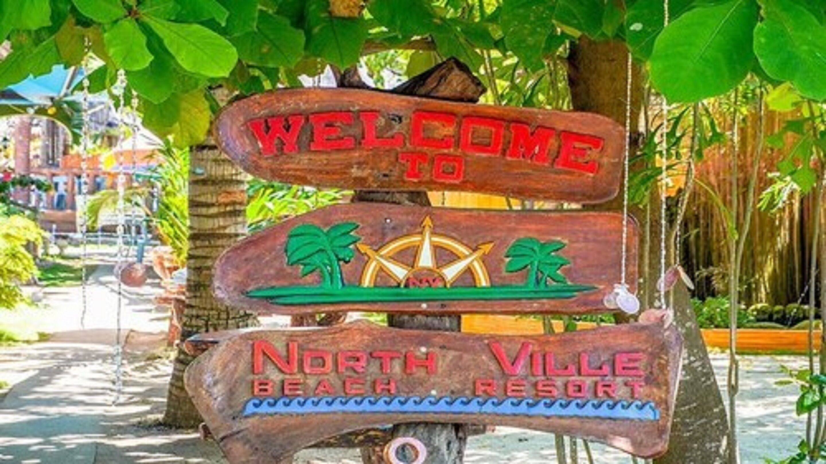 Une pancarte en bois sur laquelle est inscrit « BIENVENUE AU NORTH VILLE BEACH RESORT », avec des palmiers et une boussole.