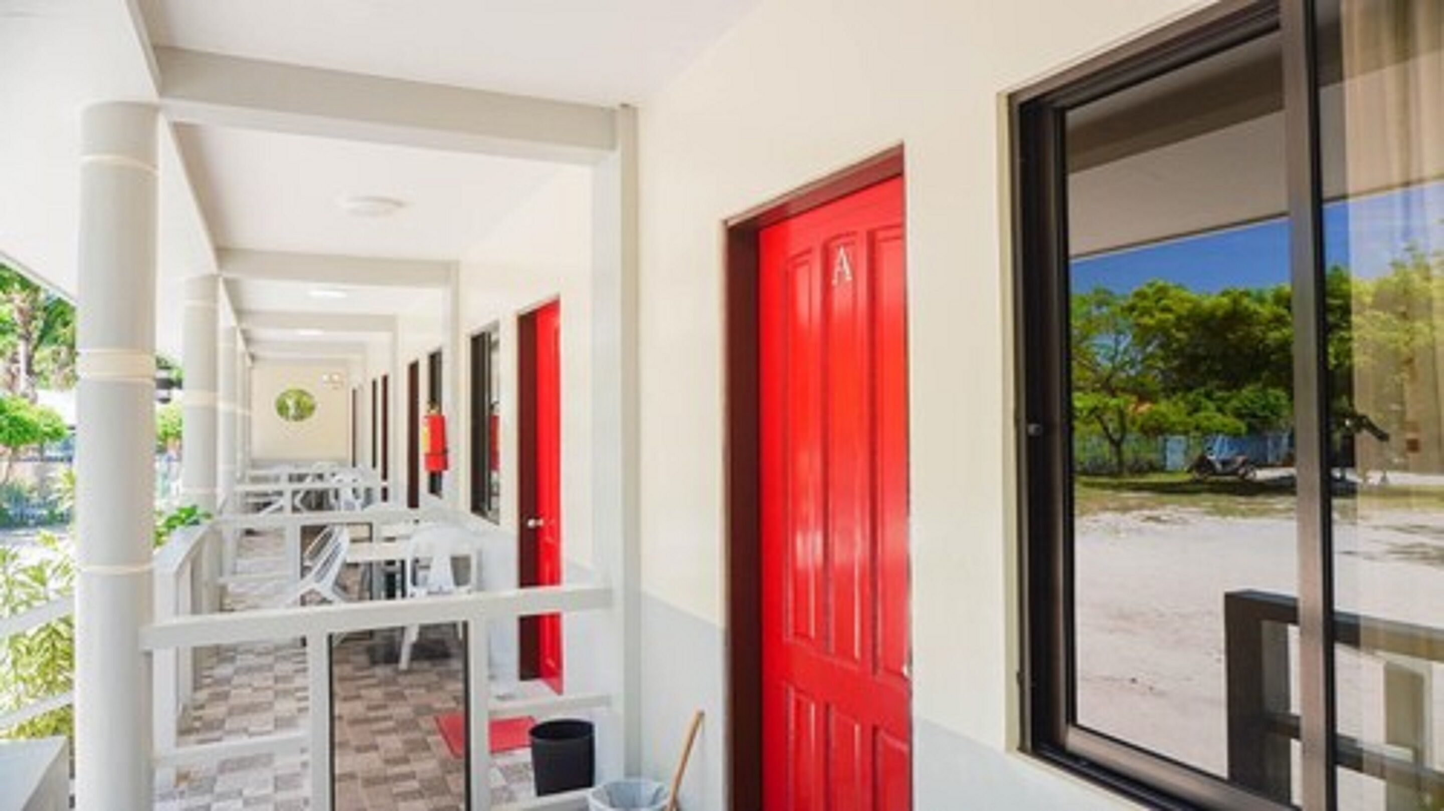 A corridor with white pillars, red doors, and a window with a view of trees and a blue sky.
