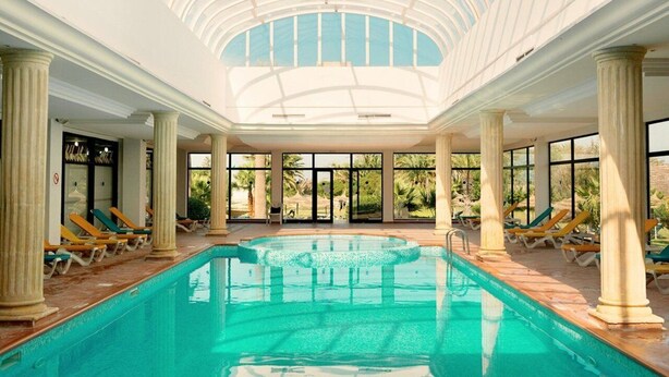 Une piscine intérieure dotée d’un toit en verre, entourée de colonnes et de chaises longues.