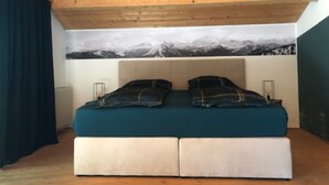 A bedroom with a wooden ceiling, a bed with a teal bedspread and plaid pillows, and a mountain landscape mural on the wall. - Ferienwohnung Hochgern (Grassau)