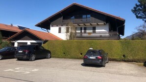 A two-story building with a balcony, parked cars, and a clear blue sky. - Ferienwohnung Hochgern (Grassau)