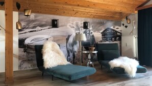 A cozy room with wooden ceiling beams, a mural of a snowy landscape, two green lounge chairs with fluffy white throws, and a table with a lamp. - Ferienwohnung Hochgern (Grassau)
