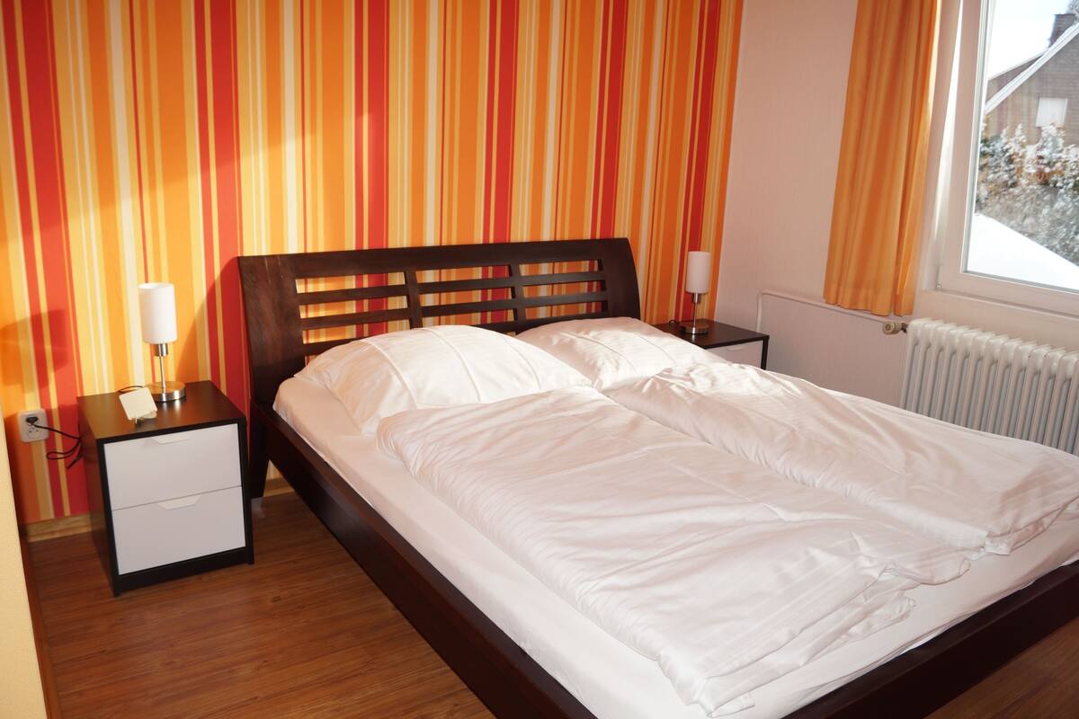 Una cama bien tendida con sábanas blancas en una habitación con papel tapiz a rayas y una ventana con cortinas.