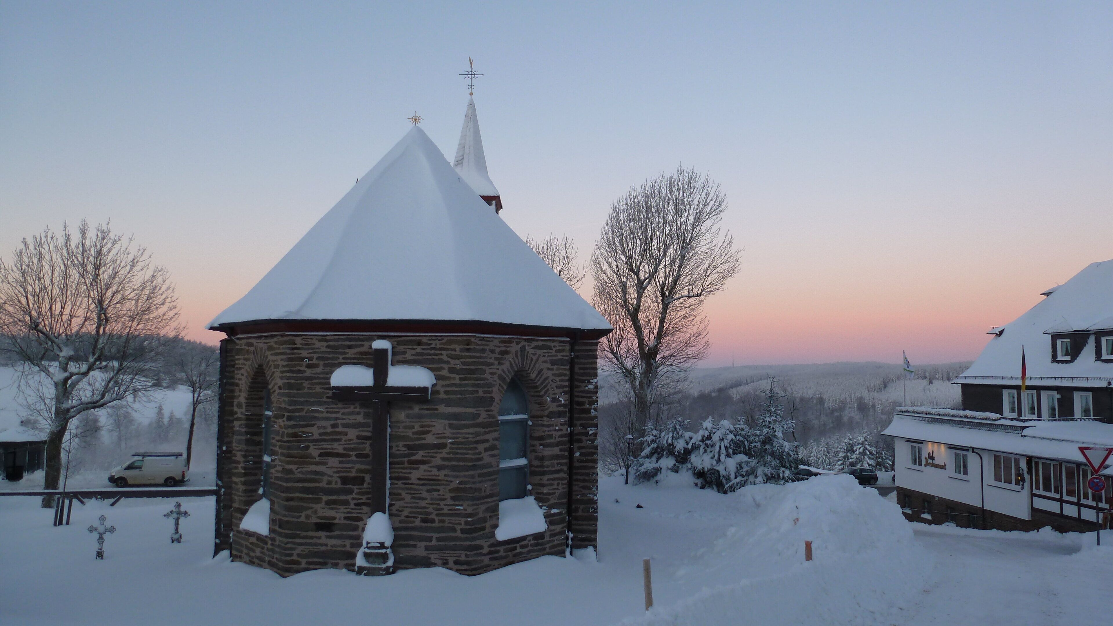 Een stenen kerk met een met sneeuw bedekte top en een klokkentoren, omringd door besneeuwde bomen en een uitzicht op een verafgelegen landschap.