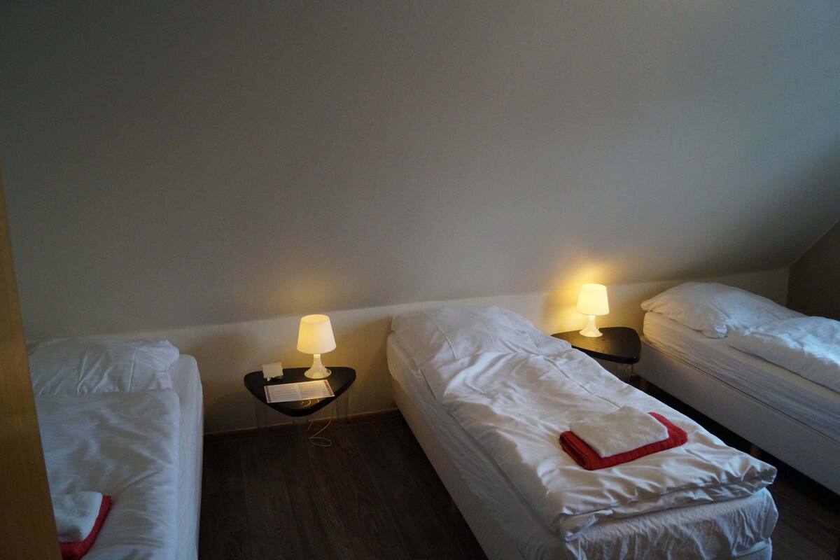 Una habitación pequeña y poco iluminada con tres camas individuales, cada una con una mesita de noche y una lámpara.