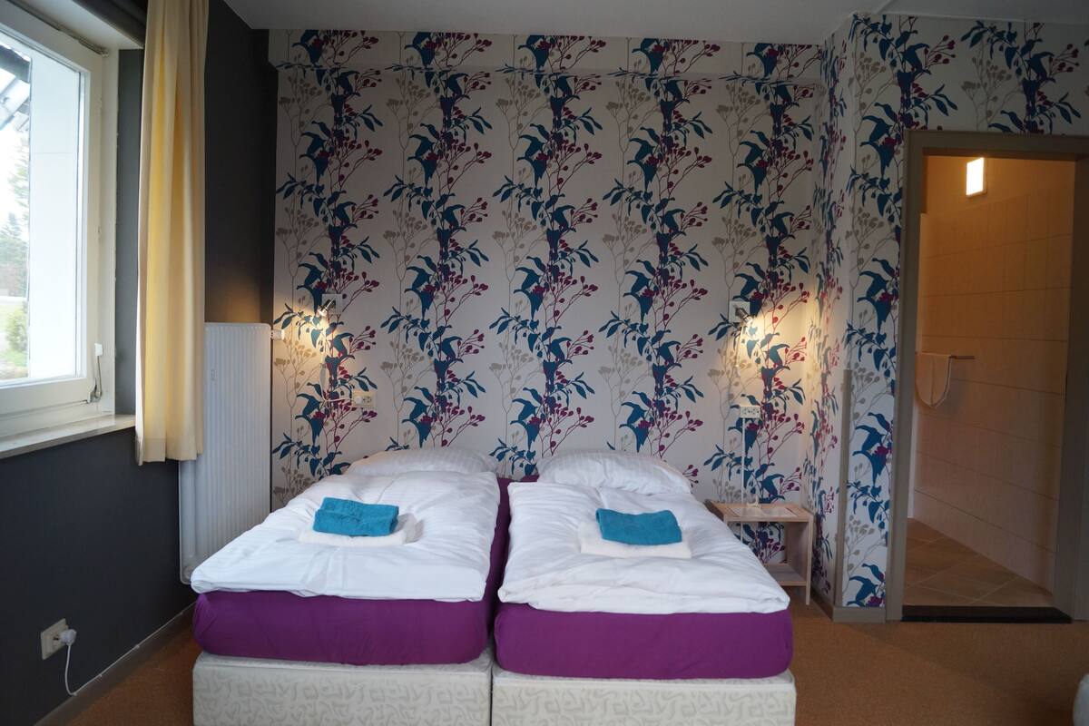 Una habitación con dos camas, papel tapiz floral, una ventana con cortinas y una puerta que da a otra habitación.