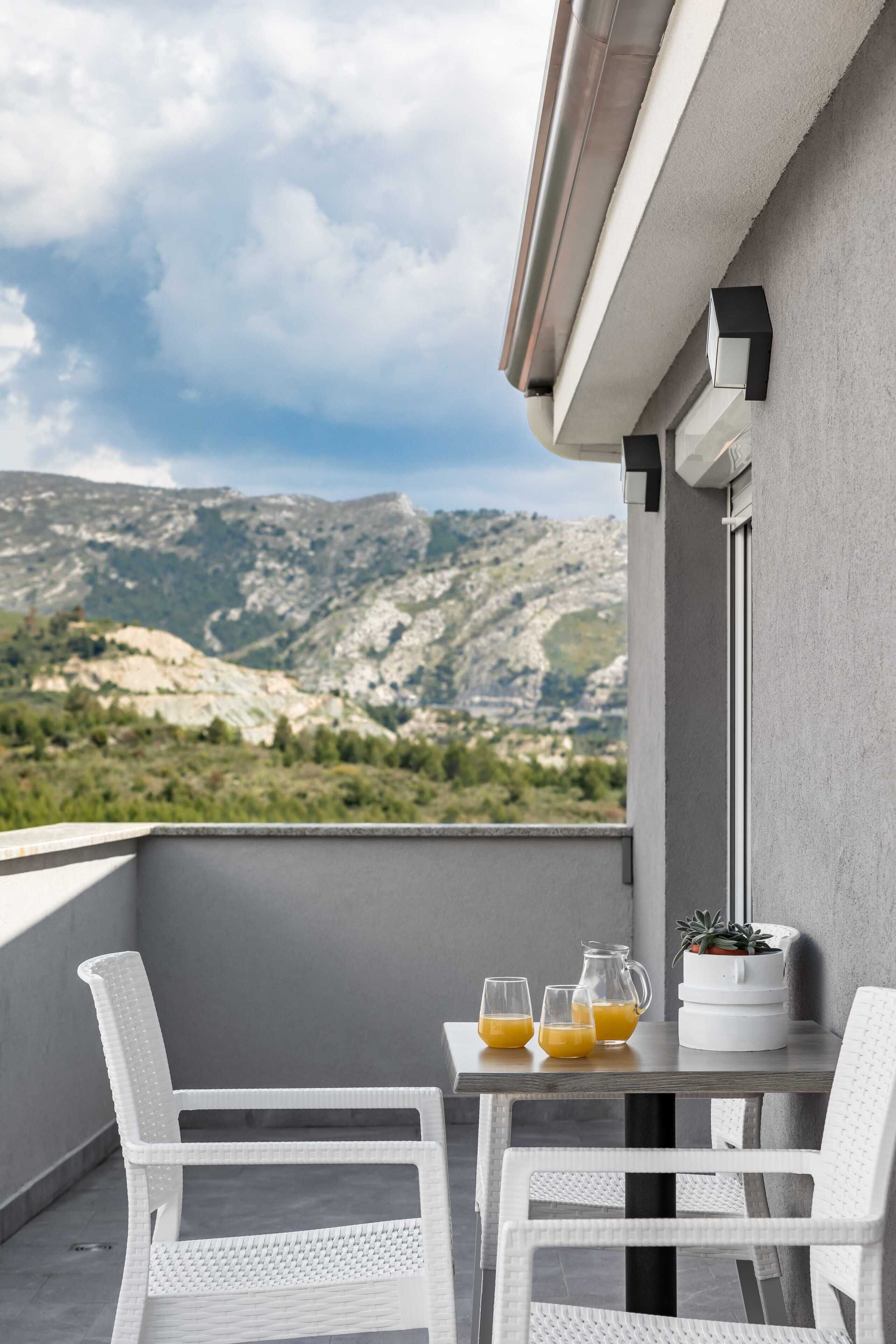 A balcony with white wicker chairs, a table with drinks, and a view of mountains.