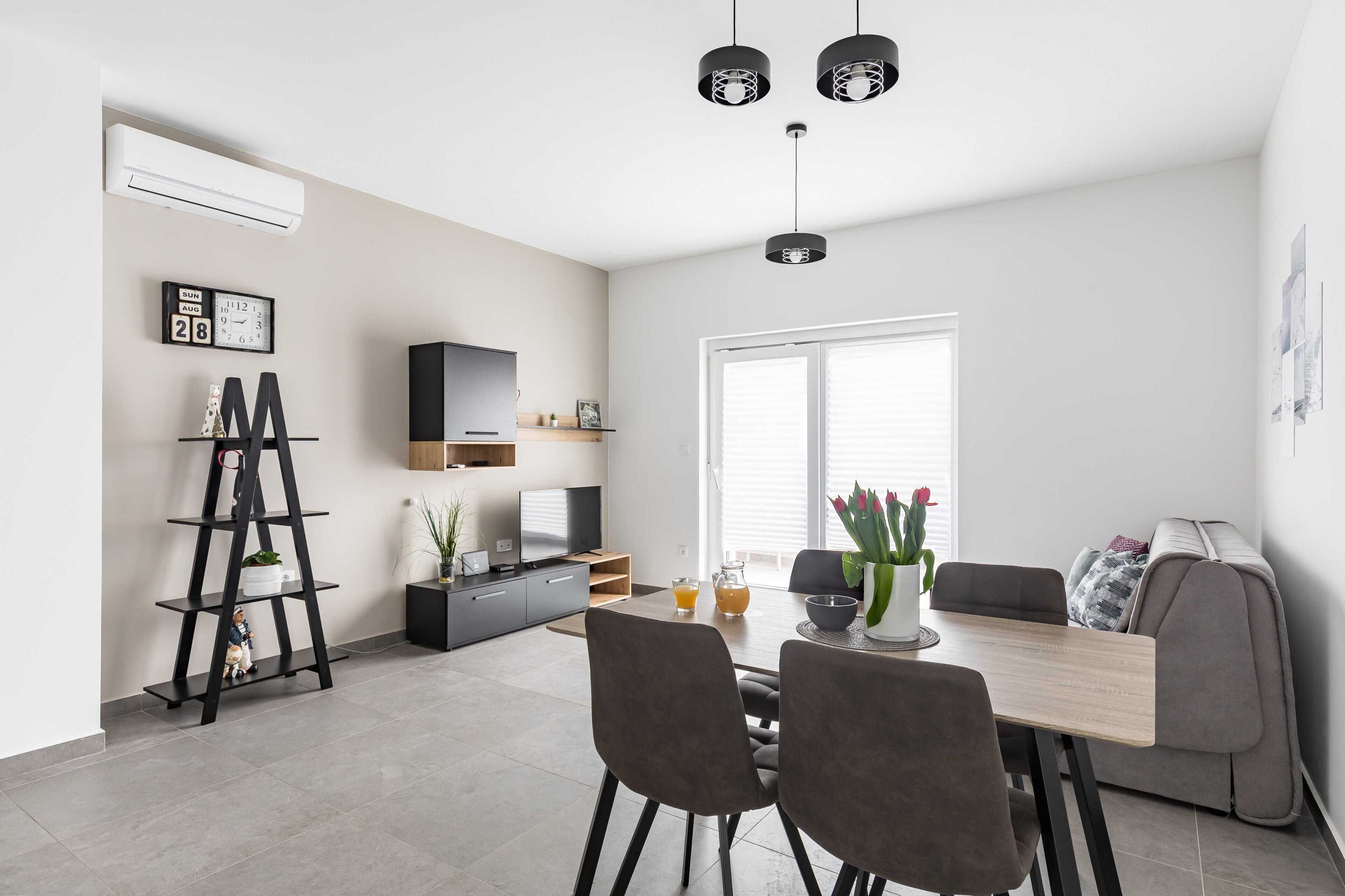 A modern dining area with a wooden table, grey chairs, and a grey sofa. There is a black shelf with decorative items and a black ladder. A television is mounted on the wall, and a window with blinds is visible.