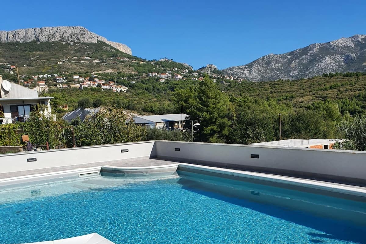 A swimming pool with clear blue water, surrounded by a tiled deck. In the background, there are houses and mountains.