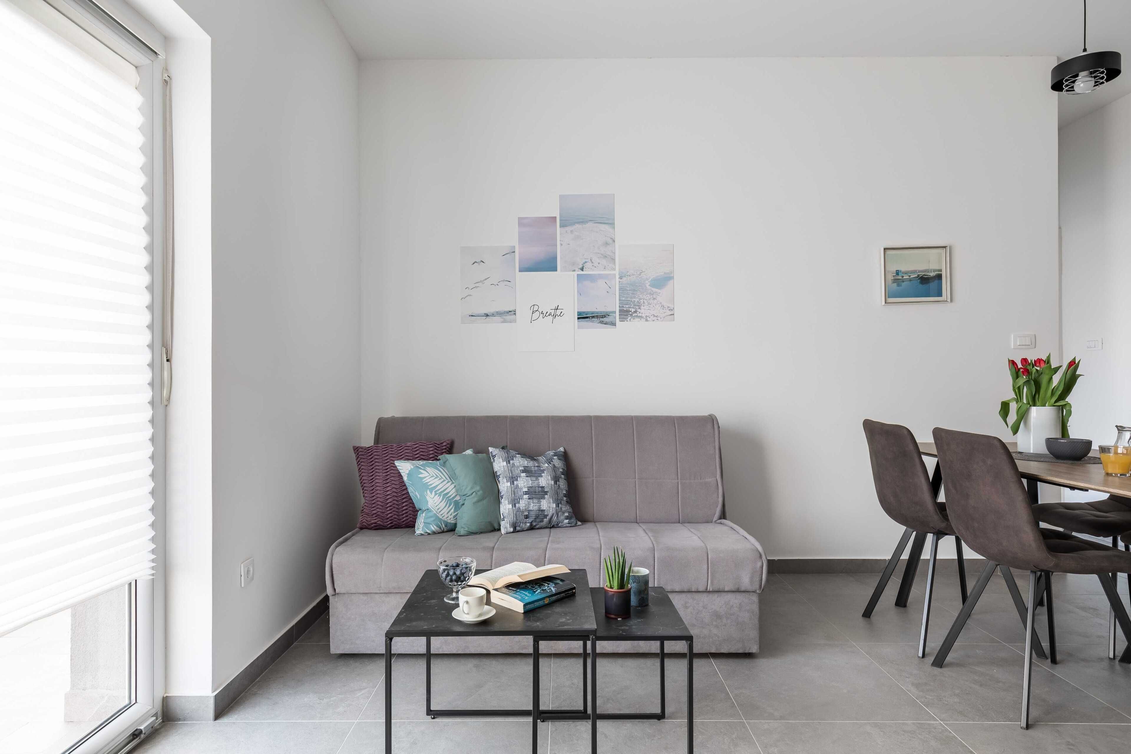 A modern living room with a grey sofa, a coffee table with books and a cup, a dining area with chairs and a table, and a wall with framed artwork.