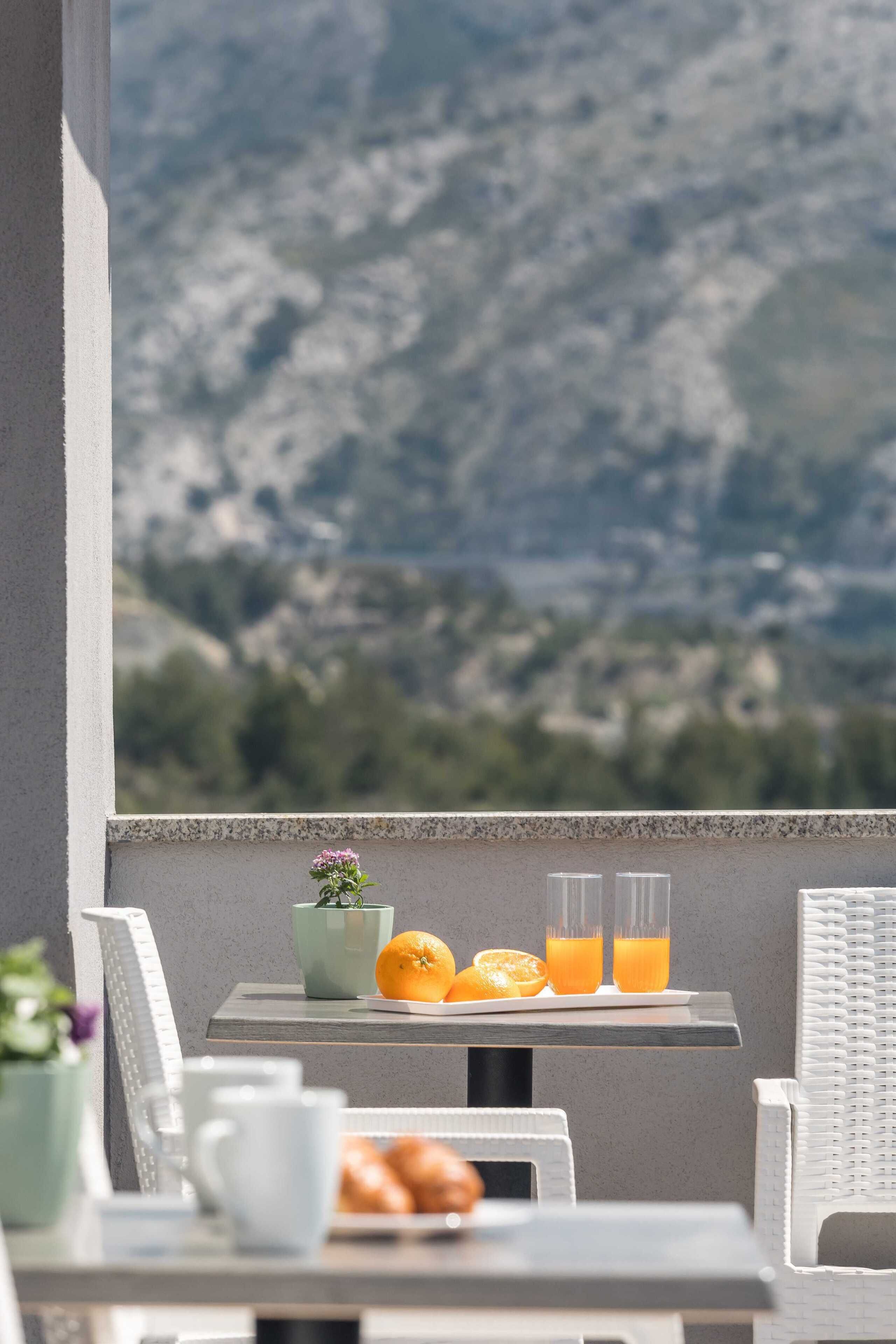 A balcony with a table set for breakfast, including oranges and orange juice, overlooking a mountainous landscape.