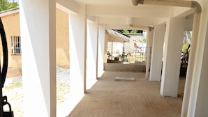 Un couloir aux murs et aux colonnes blancs, qui mène à un espace extérieur offrant une vue sur la verdure et un bâtiment en arrière-plan.