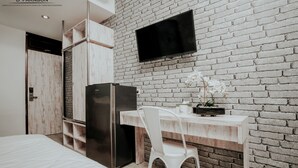 A modern room with a brick wall, a flat-screen TV, a white desk, a white chair, a black refrigerator, and a bed with a wooden headboard. - Dparagon Kemuning - Campus Accommodation (Depok)
