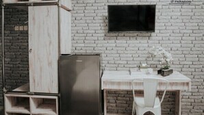A room with a white brick wall, a flat-screen TV mounted above a white desk, and a black refrigerator. - Dparagon Kemuning - Campus Accommodation (Depok)