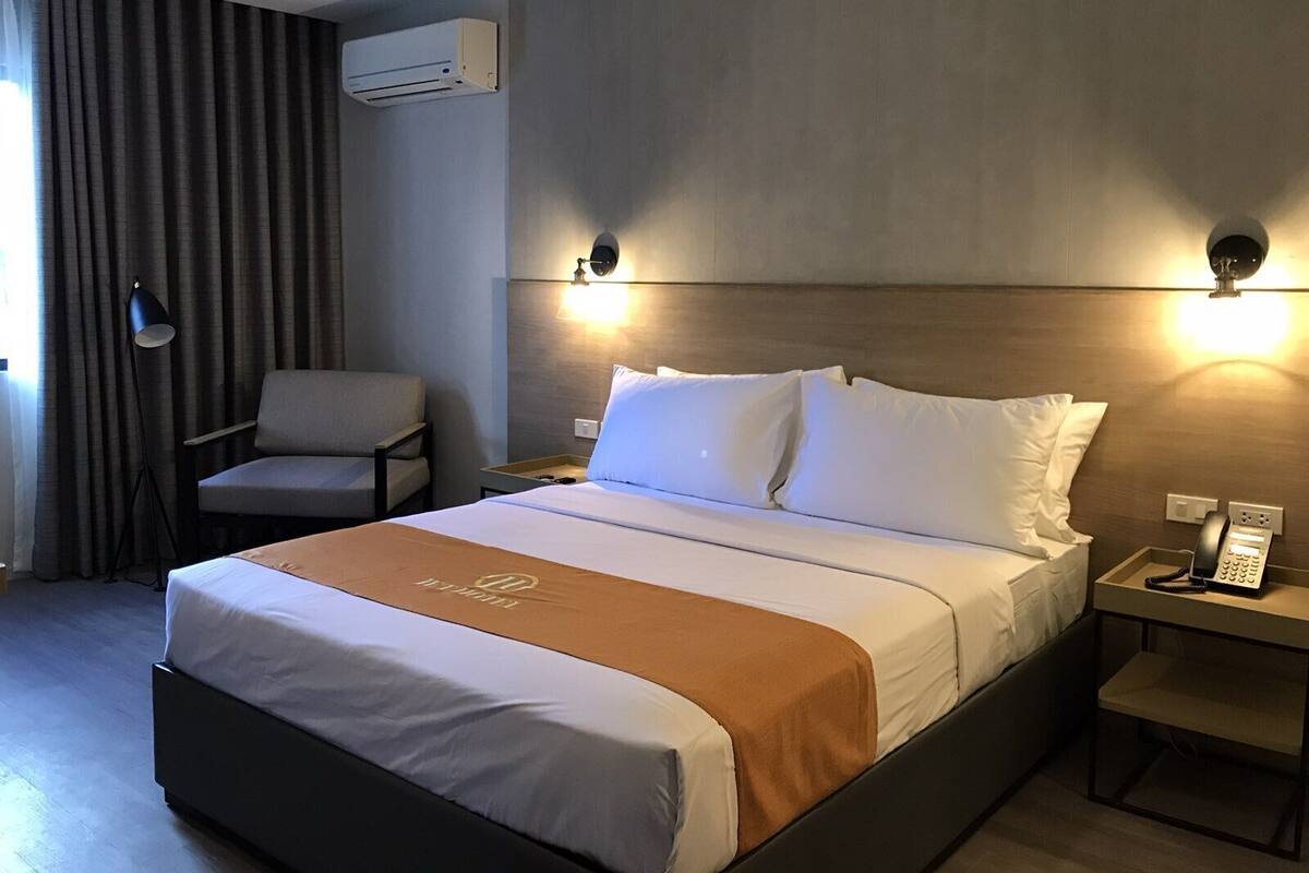 Quarto de hotel com uma cama, uma cadeira, mesa de cabeceira com telefone e ar condicionado.