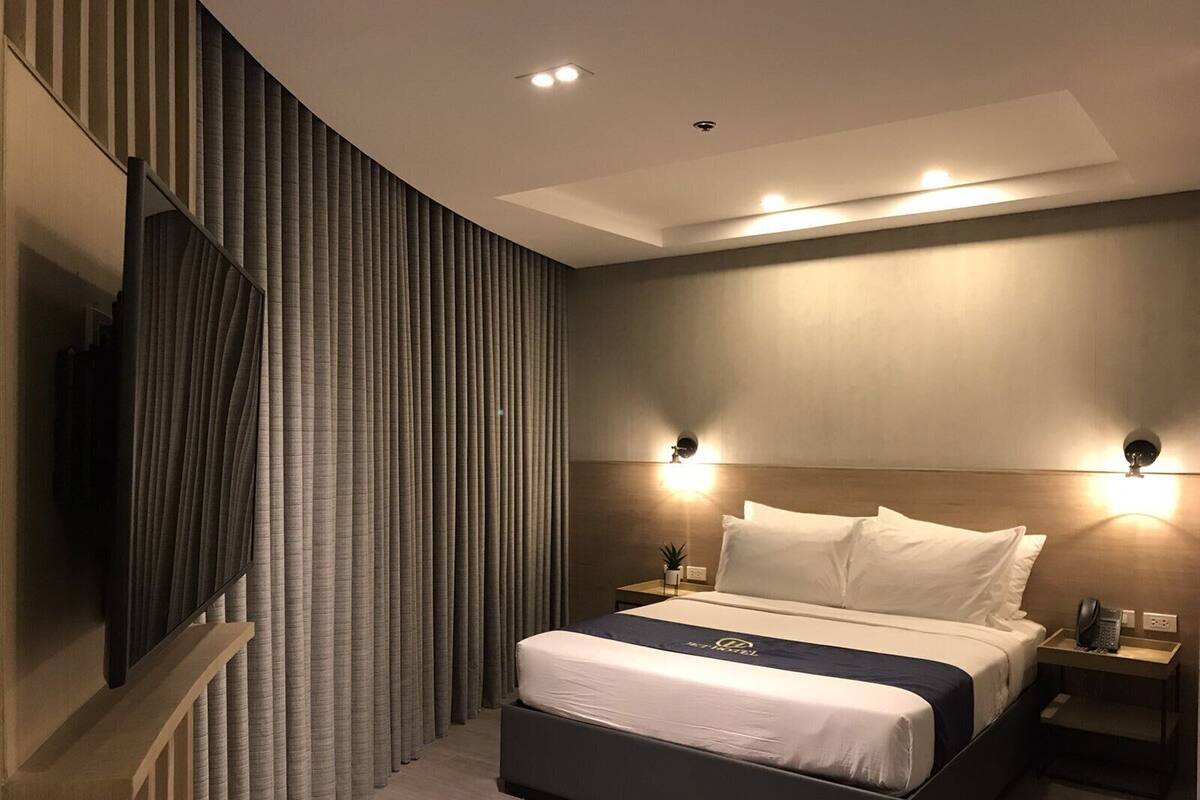 Quarto de hotel moderno com uma cama grande, duas mesas de cabeceira, uma TV fixada na parede e um lustre no teto.