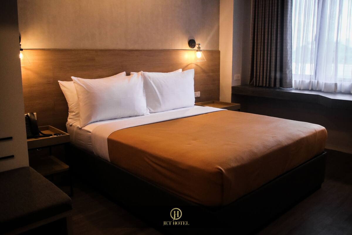 Quarto de hotel com uma cama grande, uma mesa de cabeceira e uma janela com cortinas.